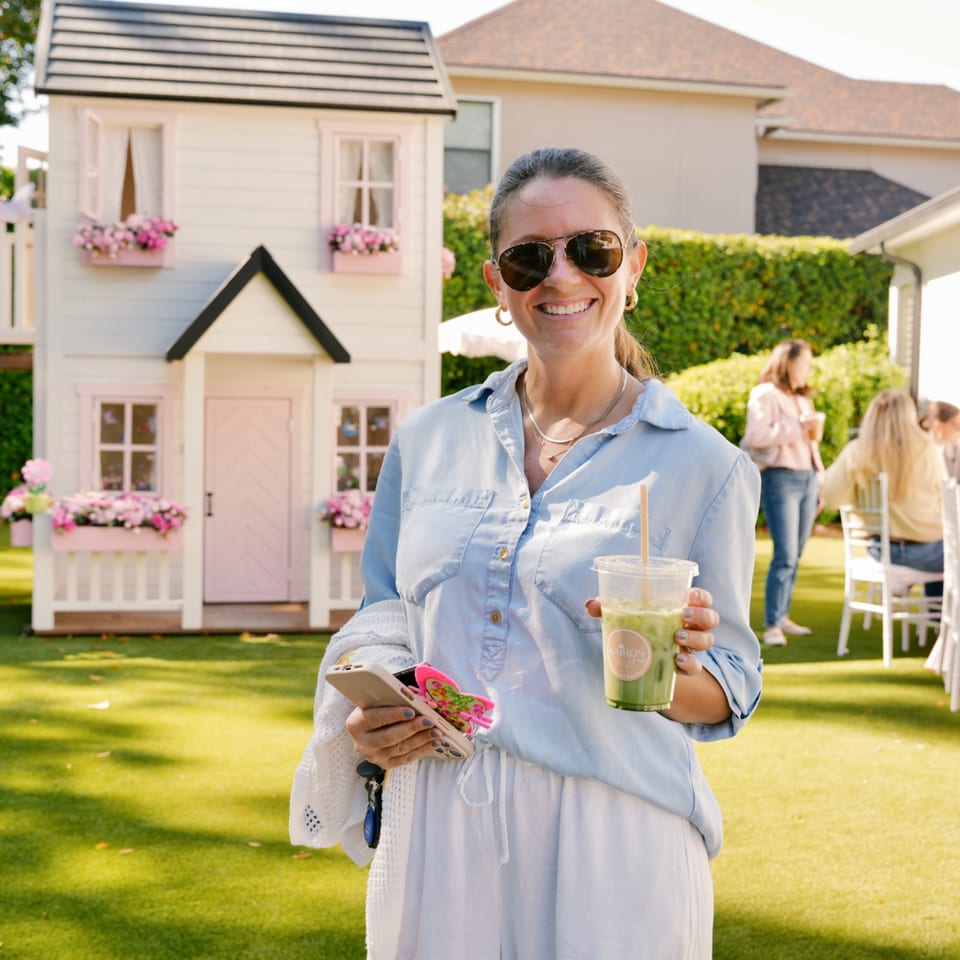 Smiling woman in sunglasses holds a Kairos Jax green drink in a sunny backyard with a playhouse.