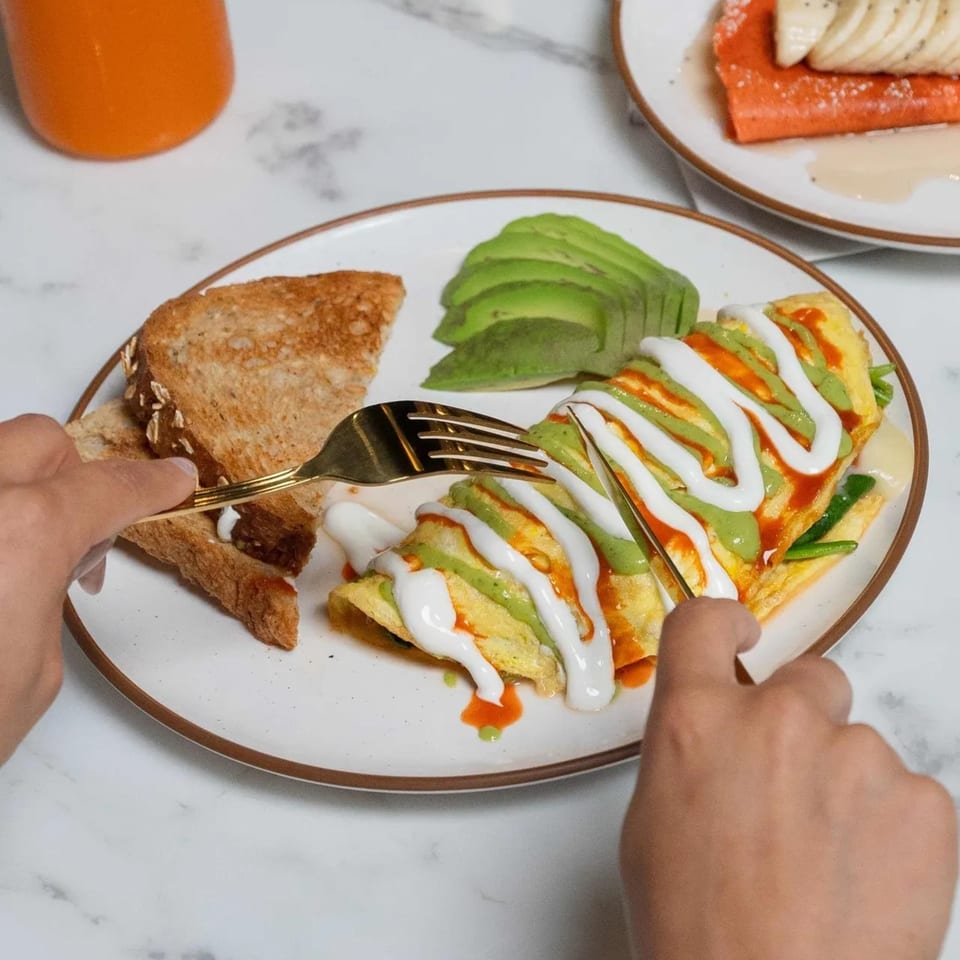 Hands cutting into a Kairos Jax omelet with avocado, toast, and sauces on a white plate.