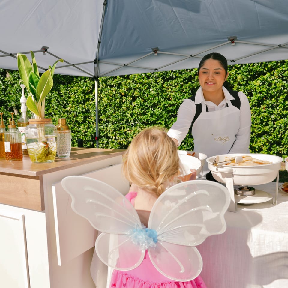 Kairos Jax staff serving food to a child in a pink dress with fairy wings under a tent.