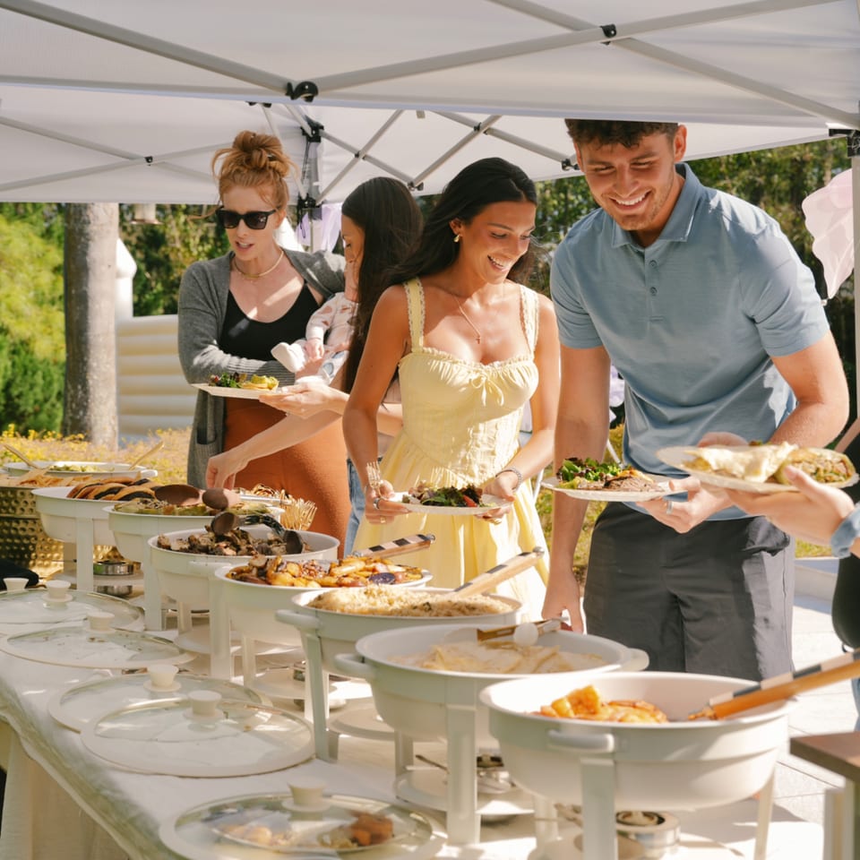 People serving themselves healthy food from a buffet line under a white tent outdoors.